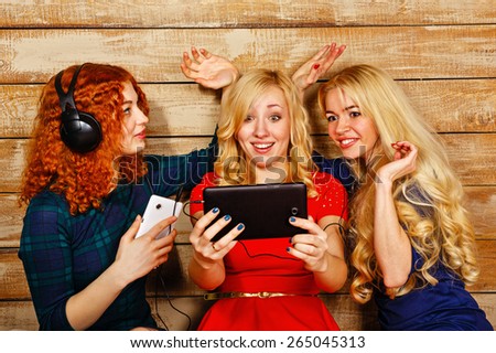 Three sisters, blond and red, listening to music on headphones. Girl doing funny ears hands sister, looking at screen the tablet PC, making happy selfie. Sisters sitting in the bedroom.