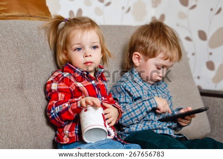 Happy brother and sister sitting on the couch. Little cute girl holding a mug in her hands, and the boy is holding a cell phone. Love, Family, Friendship