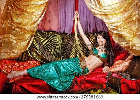Sexy oriental beauty is in the tent on the pillows and a sword in her hand rests on her hip. The concept of the Arab harem.