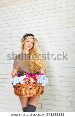 Attractive woman dressed in a sweater and cap holding a basket of flowers. Girl smiling, standing at a brick wall. The concept of innocent beauty.