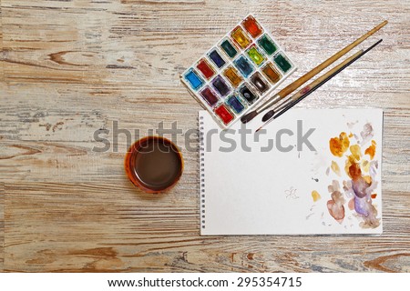 Watercolor paint, brushes and sketch pad on a wooden table. Watercolor spots on the paper. Painter Tools group.