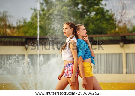 Best friends stand back to back near the fountain in summer park. Girls dressed in shorts and a shirt. On summer vacation. The concept of true friendship.