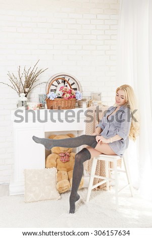 Pretty blonde girl straightens stockings sitting on a chair by the fireplace. The concept of home comfort.