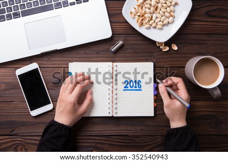 Top view office desk mockup: laptop, notebook, smartphone, snack pistachios, and cup of coffee on rustic brown wooden background. New year 2016 resolution
