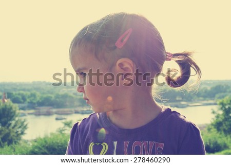 Profile of a little dark haired girl with pony tail, on a sunny day. Image filtered in faded, washed out, retro, vintage style with lens flare.