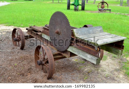 Old rusty antique mill saw with old tractor and antique wheel sitting in the background in green grassy field.