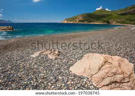 Rocks and pebble beach at Plage de Bussaglia against a turquoise sea with maquis covered rocks and clouds in background