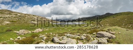 Panoramic view across Lac De Nino in Corsica with a stream meandering across a green plain in foreground and  dark clouds and snow capped mountains in the background