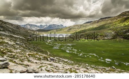 View across Lac De Nino in Corsica with a stream meandering across a green plain in foreground and  dark clouds and snow capped mountains in the background