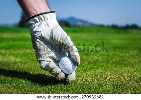 Hand with a glove is placing a tee with golf ball in the ground. Golf course with green grass with mountains in the background. Soft focus or shallow depth of field.