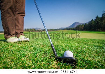 Golfer is chipping a golf ball onto the green with driver golf club. Legs and feet in the background. Green grass with forrest and mountains in the background. Soft focus or shallow depth of field.