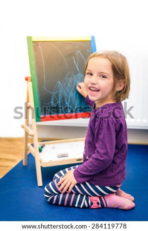 Child is drawing with pieces of color chalk on the chalk board. Girl is expressing creativity and looking at the camera, smiling in a nursery, classroom or playroom. Concept of expression and learning