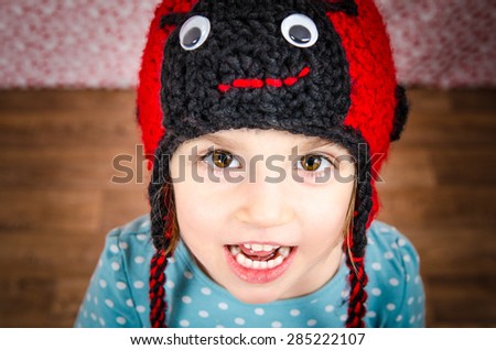 Portrait of little girl smiling and being happy, looking in the camera with big brown eyes. Child is wearing a woolen cap in the shape of ladybird on a background with red dots and wooden floor. Happy