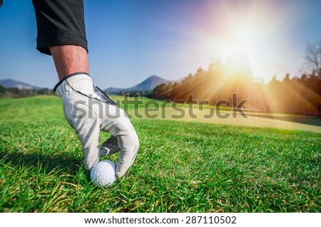 Hand with a glove is placing a t golf ball on the ground. Golf course with green grass with mountains in the background. Soft focus or shallow depth of field. Sunshine in the background