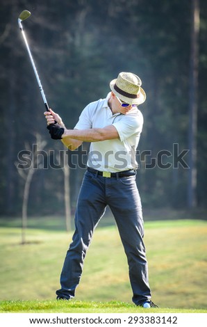 Man playing golf is swinging the golf club. He is wearing a white polo shirt, sunglasses and a hat. He is standing on green lawn.