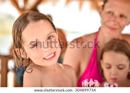 Happy family - little girl smiling and eating ice cream and mother and sister in the background. They are laughing and eating ice cream in beach hut after swimming in tropical sea. Vacation, lifestyle