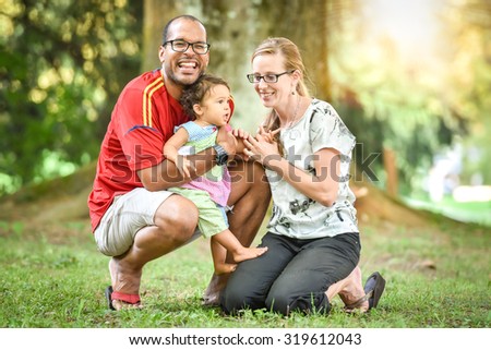 Happy interracial family is enjoying a day in the park. Little mulatto baby girl. Successful adoption. Diverse family in nature with sun in the back.