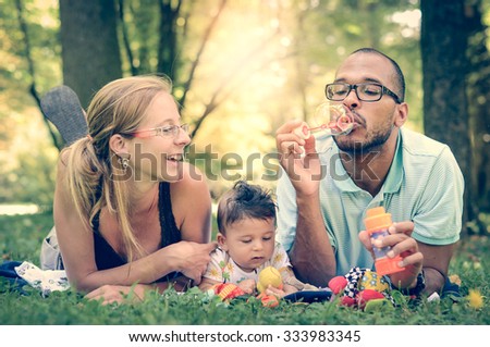 Happy interracial family blowing bubbles retro filter effect instagram filter. Mixed diverse family is enjoying a day in the park. Mother father and mulatto son smiling and picnicking in green park.