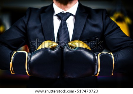 Businessman with boxing gloves is ready for corporate battle. Man in suit, shirt and a tie is holding combat gloves together. Shot in a boxing gym, concept of relentless struggle and success.