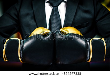 Businessman with boxing gloves is ready for corporate battle. Man in suit, shirt and a tie is holding combat gloves together. Shot in a boxing gym, concept of relentless struggle and success.