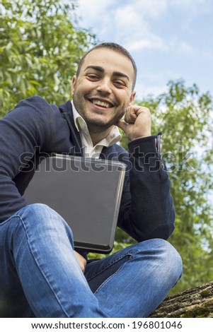 Smiling young businessman using a laptop pc computer while sitting on a tree outdoors