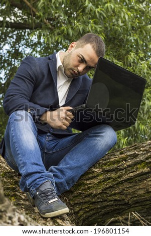 Young businessman using a laptop pc computer while sitting on a tree outdoors.