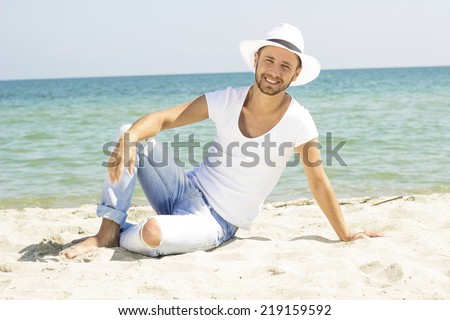 Man on beach lying in sand looking to side smiling happy wearing hipster summer hat. Young male model enjoying summer travel holiday by the ocean.