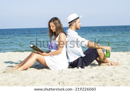 Young couple sitting in the sand on the beach leaning on each other. A woman reads a book a man drinking beer.