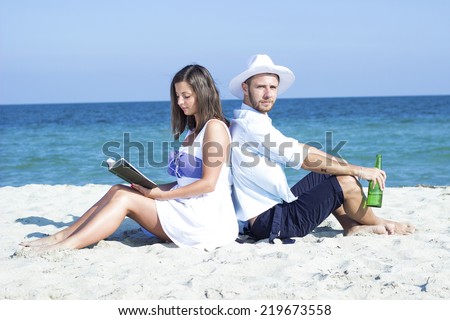 Young couple sitting in the sand on the beach leaning on each other. A woman reads a book a man drinking beer.