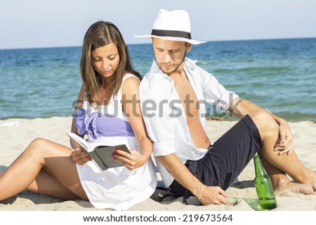 Young couple sitting in the sand on the beach leaning on each other. Women and men together read the book