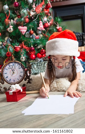 Funny girl in Santa hat writes letter to Santa near christmas tree and clock