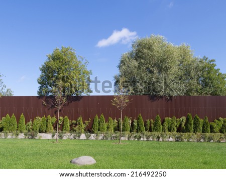 brown fence, stone on a green lawn, blue sky and trees in summer