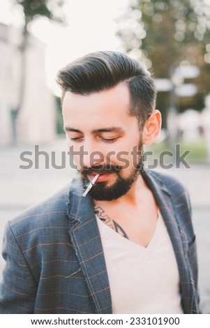 a young guy with a beard and glasses smokes a cigarette on the street