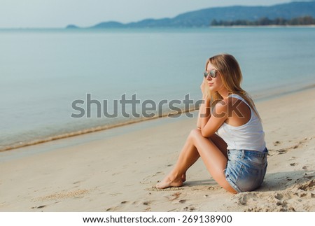 pretty girl in shorts and t-shirt sitting on the beach in sunglasses and look at the water