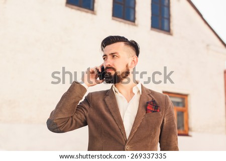 brutal man with a mustache and beard with a fashionable haircut posing on the street in a brown suit and talking on the phone