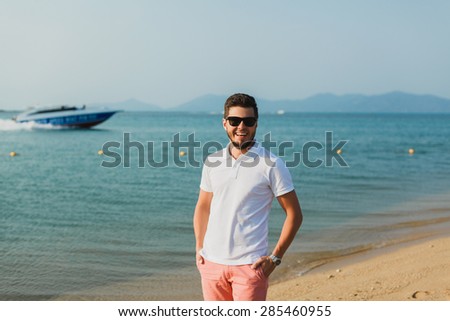 young guy in a white t-shirt and sunglasses posing on the beach and smiling and behind him floats the boat