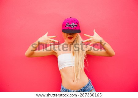 beautiful young girl in shorts and a cap poses next to a pink wall