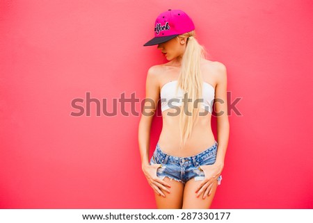 beautiful young girl in denim shorts and a cap poses next to a pink wall