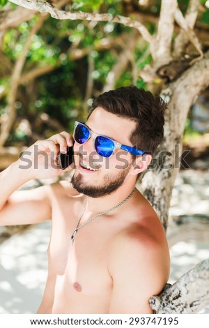 young man in sunglasses talking on the phone at the beach in a speedo