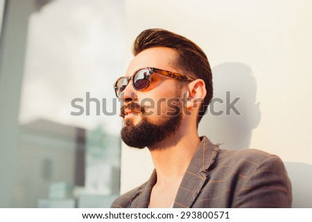 young guy with a beard and mustache with sunglasses and jacket posing on the street the sun shines