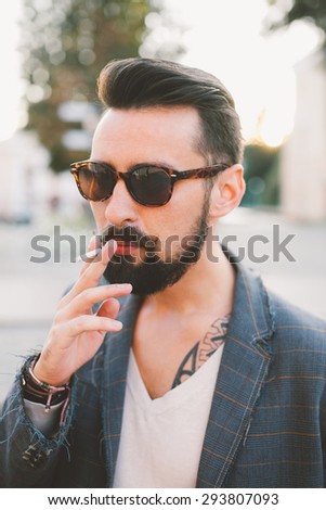 young guy with a beard and a mustache in a suit posing on the street in the sunlight and smoke a cigarett