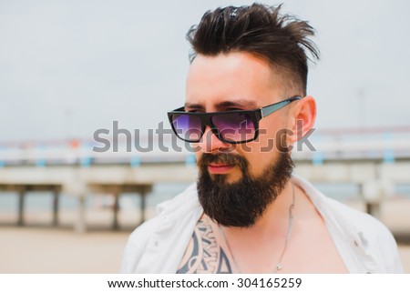 Outdoor lifestyle coolest portrait of brutal,tattooed man,posing at California beach,tattoos,denim jeans sunglasses.west coast,Florida beach,Hipster man on the beach