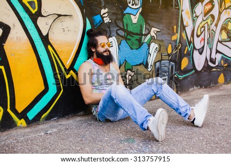 Trendy portrait of hipster  portrait.Stylish man posing with cool hairstyle,wall,ready to car trip.Happy and active.American street style,sportsman,young dj,t-shirt and shorts,Cool beard.big earphones