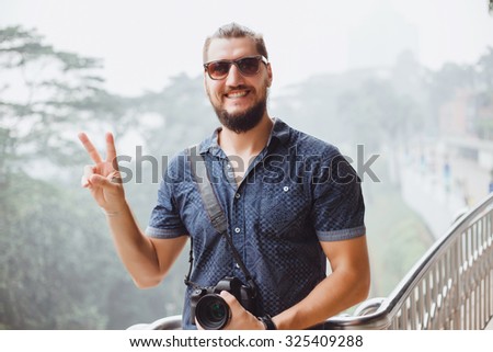lifestyle happy young guy in sunglasses and a fashionable hairstyle make professional  pictures,bearded man,hiker man,holding big camera,man with perfect white teeth,cute smile,shows peace sign,cool