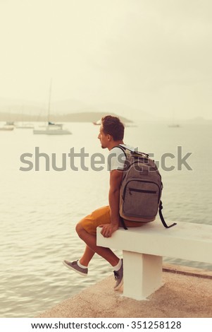 young man with beard posing in a white t-shirt standing on the pier near the sea with a backpack on shoulders
