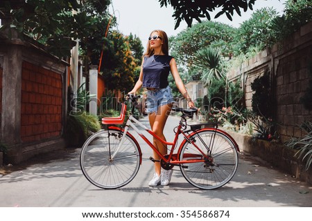 Young pretty sensual blond girl posing outdoor  with red vintage bicycle in a blue shirt in white sneakers, fashionable stylish clothes sunglasses red lipstick on her lips