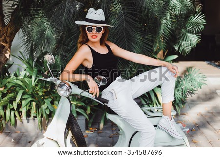 young girl in a straw hat white shirt black jeans and sneakers posing on a vintage scooter near green palms hotel in Italy  stylish girl in fashionable clothes sunglasses and red lipstick on her lips