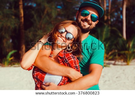 beautiful young couple of hipsters in trendy clothes, the bearded guy, t-shirt, shorts, blue baseball cap, wearing sunglasses, smiling, happy lovers,at the beach near the sea outdoor portrait,close up