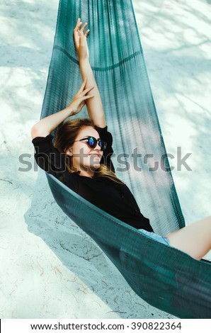 young beautiful girl in sunglasses denim shorts and shirt posing while lying on a hammock, beautiful woman, brunette resting on vacation at the beach, relax, outdoor portrait