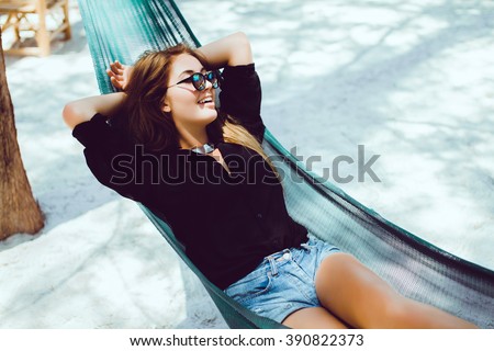 young beautiful girl in sunglasses denim shorts and shirt posing while lying on a hammock, beautiful woman, brunette resting on vacation at the beach, relax, outdoor portrait